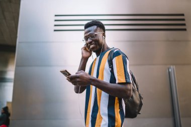 Klasik gözlüklü ve elektronik kulaklıklı komik Afrikalı Amerikalı blogcu modern akıllı telefon teknolojisinden müzik listesi seçiyor, gülümseyen hipster adam pozitif şarkıdan ve çevrimiçi sohbetten hoşlanıyor.