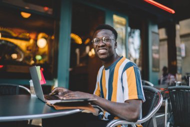 Klasik gözlüklü neşeli erkek programcının portresi kameraya gülümseyen, serbest yaşam tarzının keyfini çıkaran, dizüstü bilgisayarlı mutlu Afrikalı Amerikalı hippi çocuk.