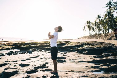 Aktif kıyafet giymiş çıplak ayaklı bir erkeğin sahilin ıslak kumlarında su ve tropikal ağaçlara karşı tek başına poz verişi.