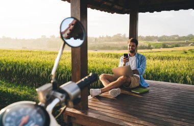 Tam vücut, sakallı, sakallı orta yaşlı bir erkek bacak bacak üstüne atmış, akıllı telefon kullanıyor ve otlağın yakınındaki motosikletli bir taraçada bilgisayarla çalışıyor.