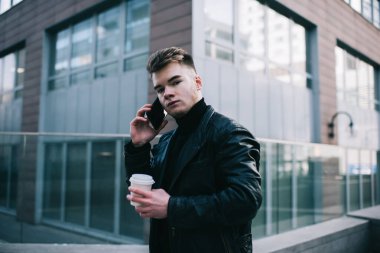 Siyah balıkçı yaka ve deri ceketli genç şık bir adam telefonda konuşuyor ve kameraya bakıyor caddede mola verip elinde bir bardak içki tutarken.