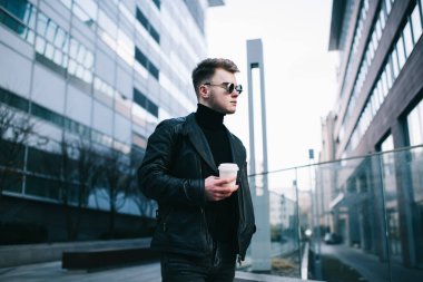 Siyah elbiseler ve yuvarlak gözlükler takan genç ve şık erkek girişimcilerin yan görüntüsü. Elinde içecek dolu bir bardak var ve dinlenirken gözlerini kaçırıyor.