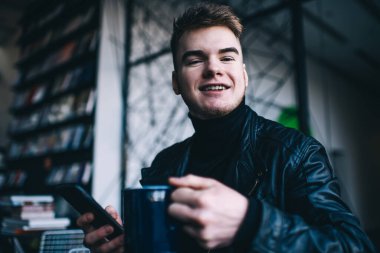 Çağdaş kitapçıda kameraya bakıp gülümserken cep telefonu kullanan bir kupa sıcak aromatik içecekle yetinen genç bir erkek.