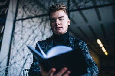 Düşük açılı, kahverengi saçlı, siyah balıkçı yaka kazaklı, deri ceketli, modern çalışma alanında kitap okuyan genç bir erkek.