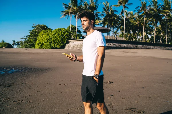 朝の海を楽しみながら海岸に一人で立っている間 携帯電話を手にスポーツウェアの若い男性選手 — ストック写真