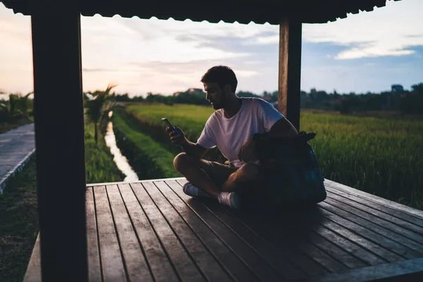 年轻的男性旅行者 背着背包坐在绿地附近的木制Pergola中 在温暖的昏暗的夜晚用智能手机使用旅游导航应用程序 — 图库照片