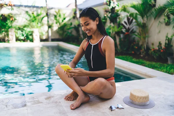 Ethnic Female Toothy Smile Swimsuit Sitting Crossed Legs Using Smartphone — Stockfoto