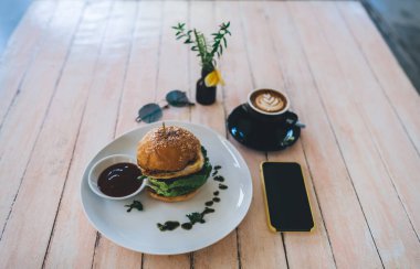 İştah açıcı taze burger ve bir fincan kahve. Akıllı telefonun yanına koyulmuş. Açık kafedeki siyah ekranlı.