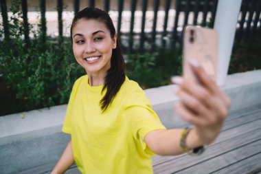 Sarı tişörtlü mutlu bir Asyalı kadın. Geniş gülümsüyor ve yazın cep telefonuyla selfie çekiyor.