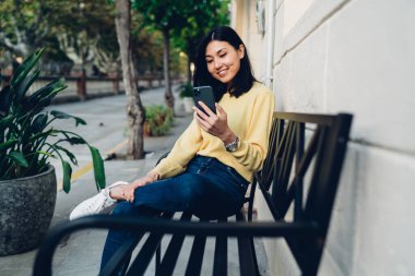 Metal bankta oturup gülümseyen ve sokakta akıllı telefonları karıştıran sıradan giysili neşeli Asyalı kadın.