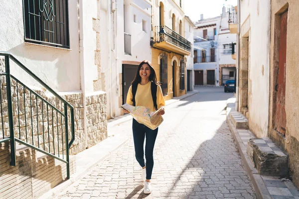 Gündelik giysiler ve sırt çantalı, elinde harita olan ve kameraya bakarken kaldırımda yürüyen neşeli etnik bir kadın vücudu.
