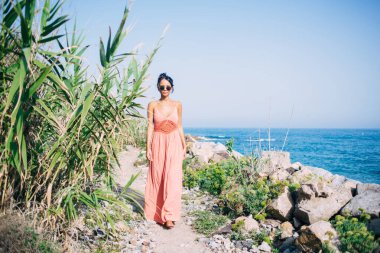 Kıyı şeridinde yürüyen ve kameraya bakan modaya uygun giyinmiş Latin turistin tam boy portresi, seyahat sırasında Filipinleri keşfeden güneş gözlüklü çekici kadın yalnız gezgin.