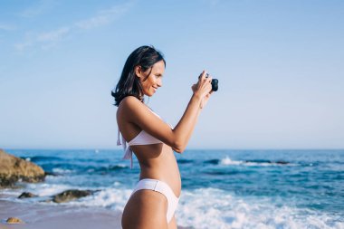 Bikini mayo giymiş neşeli Latin kadın fotoğraf hobisinin keyfini çıkarırken klasik fotoğraf makinesini test ediyor ve okyanus ufkunun görüntülerini çekiyor. Beyaz bikinili mutlu kadın yüzücü retro teknoloji kullanıyor.