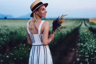 Büyük bir çiçek tarlasında dikilmiş, lavanta dalını tutarken gündelik elbise ve hasır şapkalı neşeli genç bayanın arkası.
