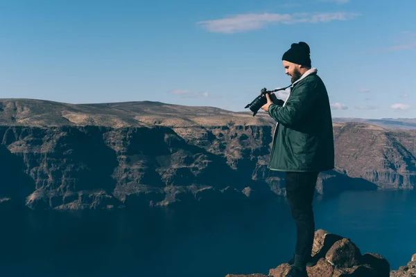 緑のジャケットと黒の帽子の穏やかな男性のフルボディは 米国全土のロードトリップ中に山とブラウジングカメラの端に立っています — ストック写真