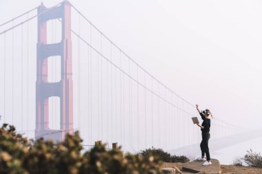 Sanal gerçeklik gözlüklü, elinde tabletle taşın üzerinde duran sisli San Francisco 'da aletleri test eden anonim kadının yan görüntüsü.
