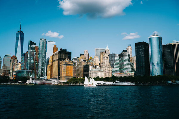 Modern tall skyscrapers with glass windows located near river with sailboat against cloudy blue sky in New York City district