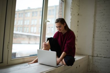Rahatlamış çıplak ayaklı kadın aydınlık odada beyaz pencere eşiğinde dinleniyor ve netbook okuyor gülümseyerek evde tek başına vakit geçiriyor.