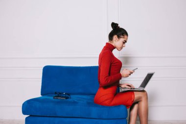Kırmızı kıyafetli genç bir kadının yan görüntüsü cep telefonundan mesaj okurken mavi koltukta otururken dizüstü bilgisayarını karıştırıyor.
