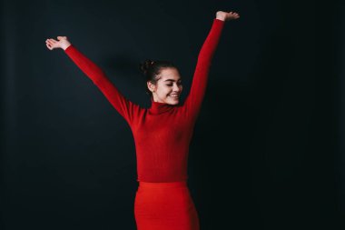 Parlak kırmızı etekli, balıkçı yakalı, gözleri kapalı ve elleri koyu gri arka planda duran neşeli genç bir kadın.