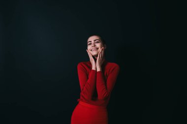 Genç, mutlu, şık renkli, dişlek gülüşlü, siyah arka planda dikilirken yukarı ve yanaklara dokunan bir model.