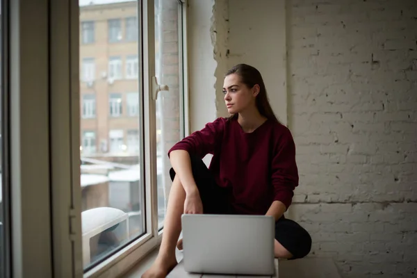 穿着休闲装的年轻而专注的女自由撰稿人 长发朝外看 坐在窗台上 在工作时与笔记本电脑保持着舒适的姿势 — 图库照片