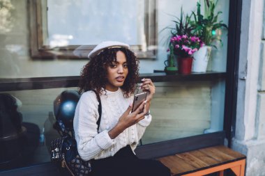Düşünceli Afro-Amerikan kadın gündelik giysiler ve şapkalı, ahşap bankta oturan ve sokakta dinlenirken akıllı telefonları karıştıran.