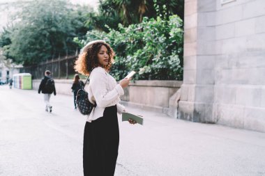 Sokakta yürürken ve akıllı telefonları karıştırırken müzik dinlerken sırt çantalı ve bereli konsantre Afro-Amerikan kadın görüntüsü