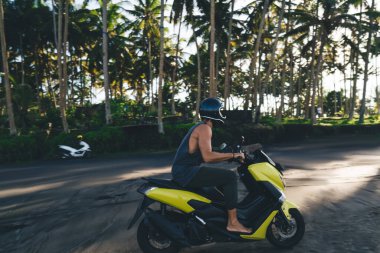Kimliği belirsiz erkek motosikletçinin koruyucu kaskı ve günlük kıyafetleri var. Motosiklet sürüyorlar. Palmiye ağaçlarının yakınındaki kumsalda.