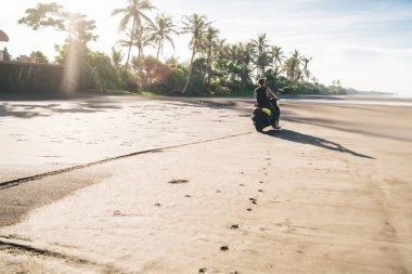 Yaz tatilinde okyanus ve palmiye ağaçları yakınlarındaki kumlu sahil boyunca motosiklet süren tanınmayan genç erkek motorcunun arka görüntüsü.