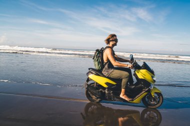 Sırt çantalı, sırt çantalı, sarı scooterlı, dalgalı, kumlu plajları yıkayan mutlu genç adamın yan görüntüsü.