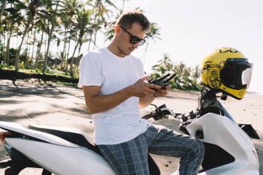 Moda kıyafetli, güneş gözlüklü, motosiklette oturan ve güneşli bir günde tropikal arka planda insansız hava aracı uzaktan kumandası kullanan genç bir gezginin yan görüntüsü.