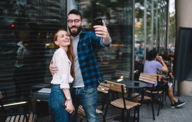 Optik gözlüklü sıradan giyinmiş sevgili cep telefonu kamerasına gülümserken aynı zamanda olumlu medya içeriği yaratıyor, aşık çift şehirde gündüz vakti video bloğu çekiyor.