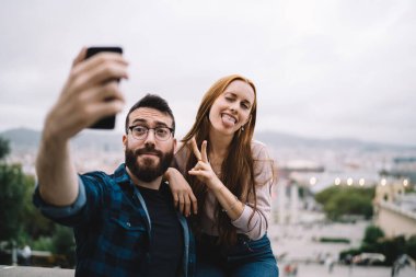 Milenyum çifti balayı sırasında fotoğraf çekmek için akıllı telefon kamerası kullanıyor, gözlüklü beyaz erkek arkadaş komik kız arkadaşıyla buluşurken mobil uygulamayla fotoğraf çekiyor.