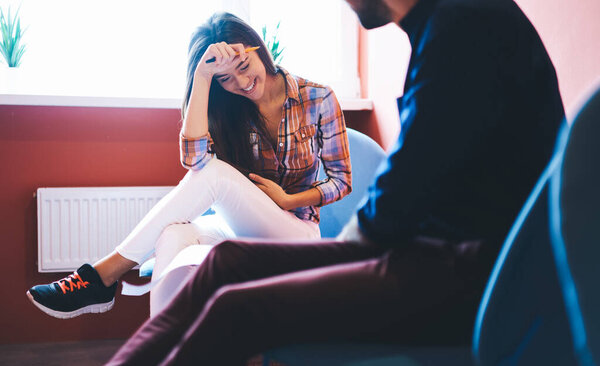 Low angle of young female in casual outfit sitting on chair and touching head while laughing having conversation with male