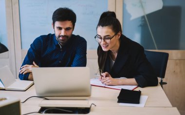 Genç gülümseyen profesyoneller dizüstü bilgisayarlarında veri analiz ediyorlar defterlere not alıyorlar ve modern ofiste masada oturmaktan memnun görünüyorlar.