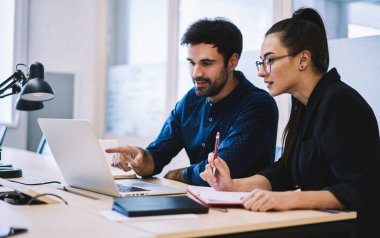 Genç, olumlu, profesyonel bir erkek, kadın meslektaşına yaptığı hatayı anlatıyor. Raporda, modern ofisteki masadaki dizüstü bilgisayarın ekranına işaret ediyor.