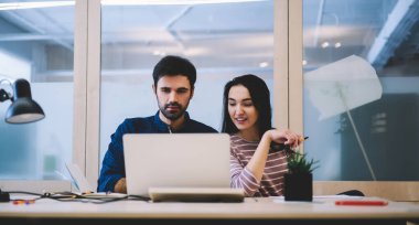 Yoğunlaştırılmış erkek ofis çalışanı dizüstü bilgisayara bakıyor ve iş projesini iş yerinde ekranlara bakarken neşeli kadın çalışanla tartışıyor.
