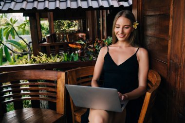 Açık kafede dizüstü bilgisayarda yazı yazan ve yaz tatilini tropikal bir ülkede tek başına geçiren pozitif kadın işçi.