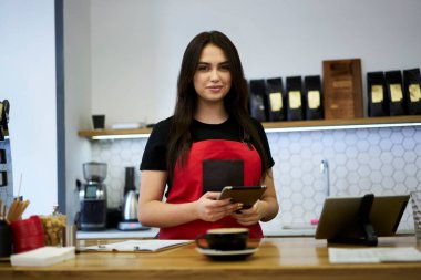 Önlüklü esmer bir kadının yarım boy portresi kafenin çalışma yerinde dijital tablet siparişini kontrol ediyor, kahve dükkanının pozitif kadın çalışanı servis için teknolojiyi kullanıyor.