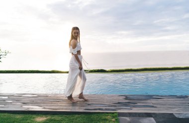 Göl kenarındaki ahşap yolda duran, beyaz elbiseli, ince, sakin bir kadının yan görüntüsü.