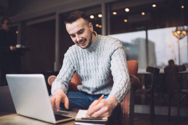 Geniş kafede çalışırken iş stratejisini kağıda yazarken bilgisayarda internette sörf yapan memnun adam.