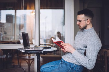 Sıcak örgü süveter ve gözlüklü genç bir erkeğin evde kitap okurken küçük bir masada oturması.