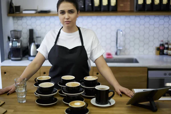お客様の注文で職場で制服姿のコーヒーハウスの白人若い女性従業員 プロの女性バリスタは カフェテリアでバーで働く多くのカプチーノカップを準備します — ストック写真