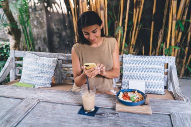Genç, etnik içerikli bir kadın cep telefonuyla mesajlaşırken sokak kafesinde lezzetli öğle yemeği ve içeceklerle dolu bir bankta oturuyor.