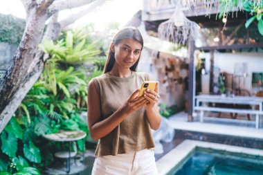 Yüzme havuzunun yanında cep telefonuyla dikilip arka taraftaki kameraya bakan gündelik kıyafetli, mutlu genç bayan.