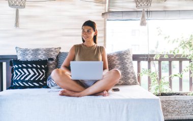 Nilüfer çiçekli elbiseli genç bir kadın güneşli bir günde Bali villasında dizüstü bilgisayar kullanarak rahat koltukta poz veriyor.