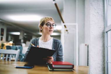 Ciddi kadın çalışanların düşük açılı günlük kıyafetleri içinde pencerenin yanındaki masaya bakar ve çalışırken notlar alır.