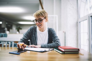 Günlük giysiler ve gözlüklerle gülümseyen kız ödevlerini yapıyor ve gündüzleri rahat kütüphanede akıllı telefon ekranlarına bakıyor.