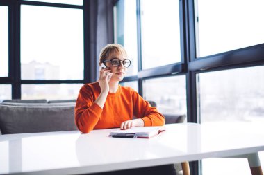 Genç, gülümseyen, turuncu kazak giyen, gözlüklü, cep telefonuyla konuşan modern salonda günlüklü bir kadın.